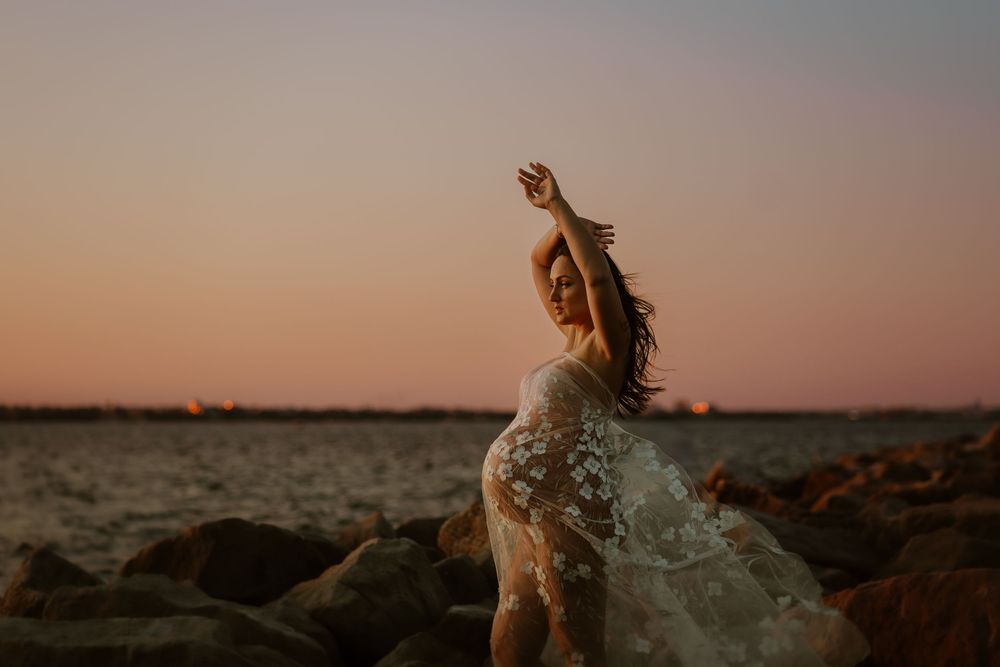 A silhouetted figure in a white dress dances on rocky shore against warm sunset sky as water splashes around them.