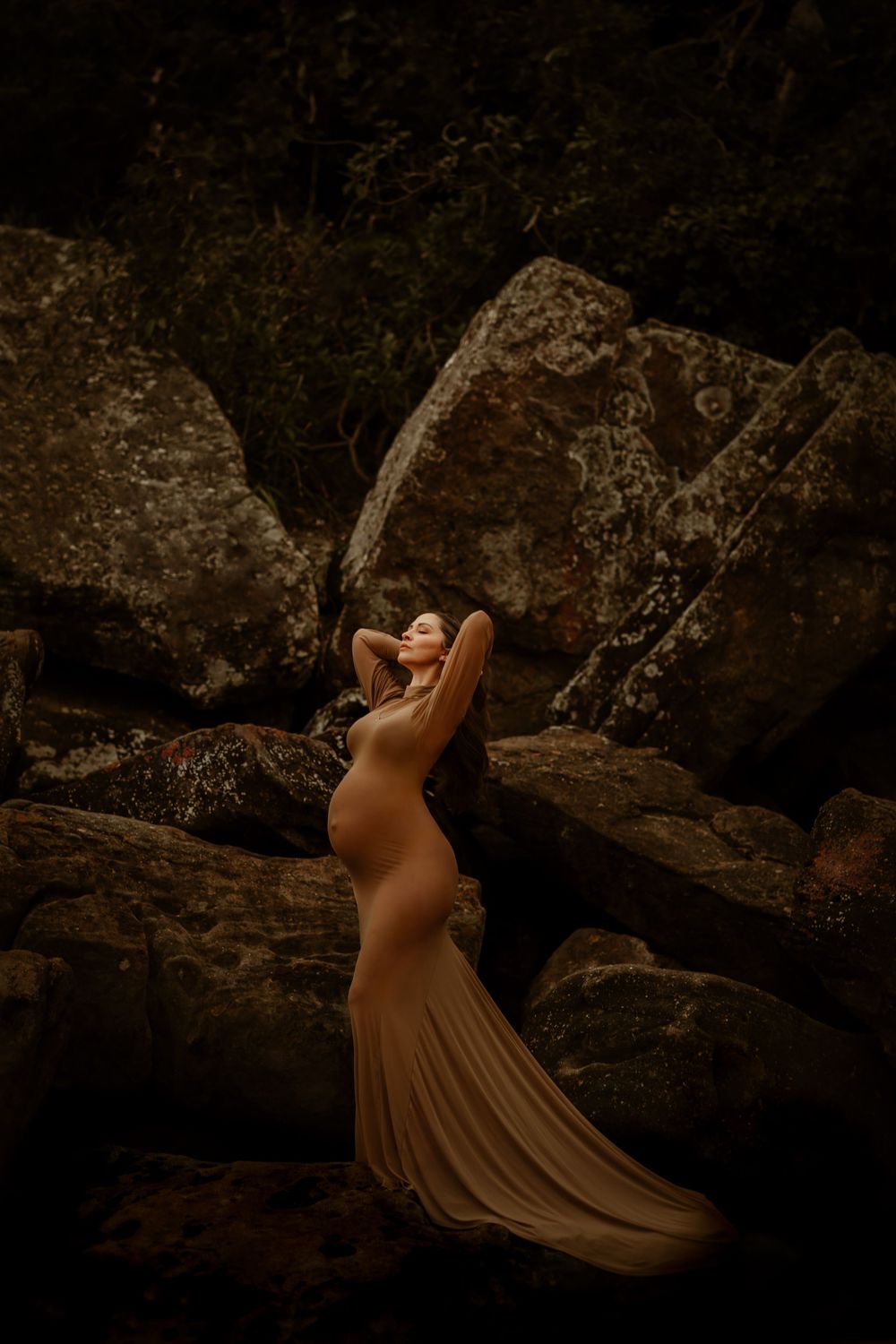 A person in a flowing brown dress poses dramatically against a dark textured cave wall or rock formation.