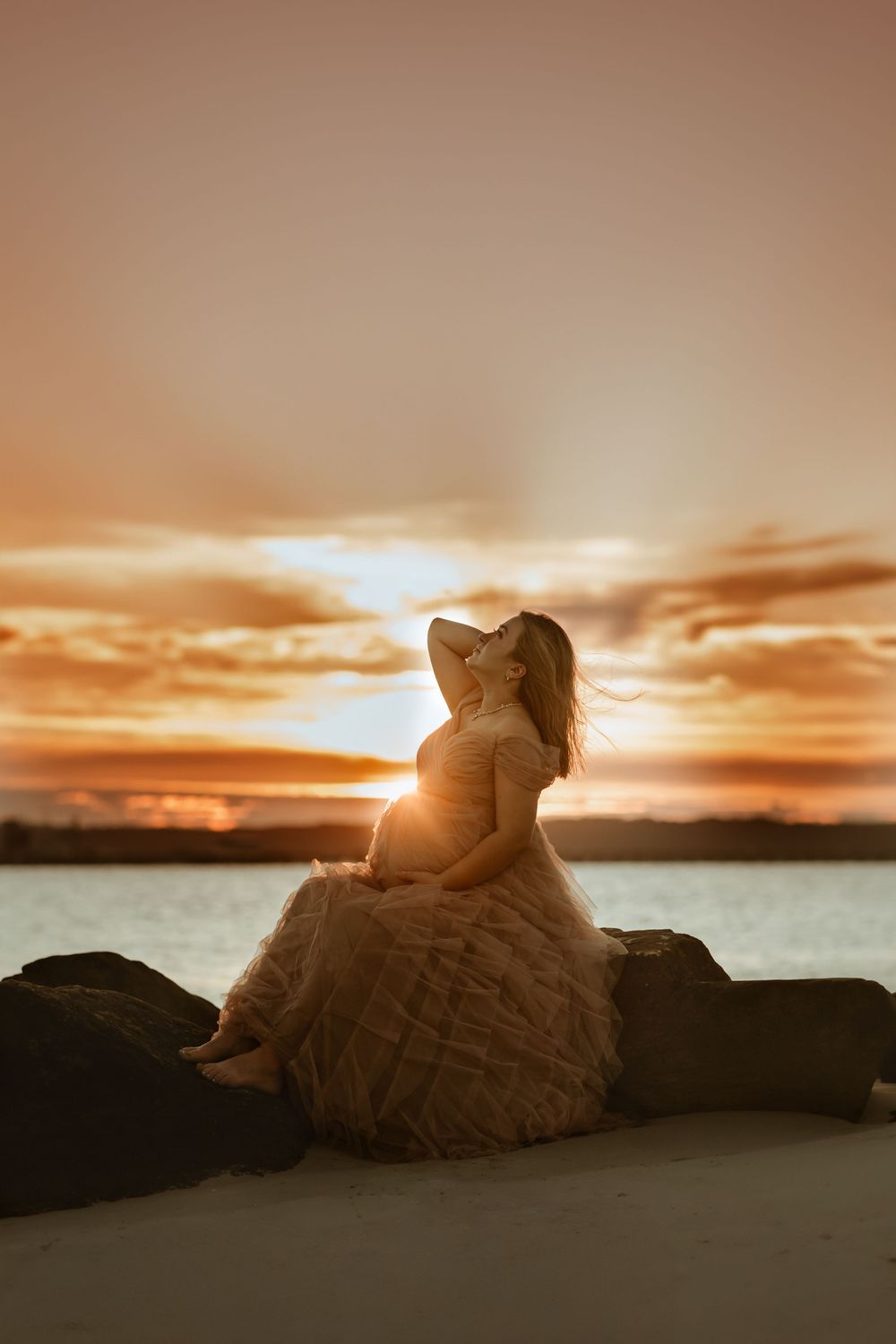 Silhouette of someone in a flowing dress sitting on rocks by the water during a dramatic golden sunset.
