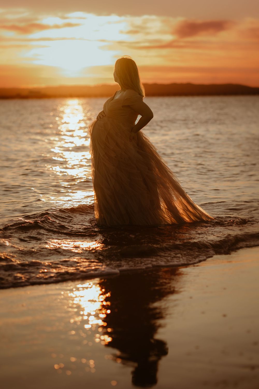 A silhouetted figure sits in shallow ocean water during a golden sunset, creating a serene and reflective scene.