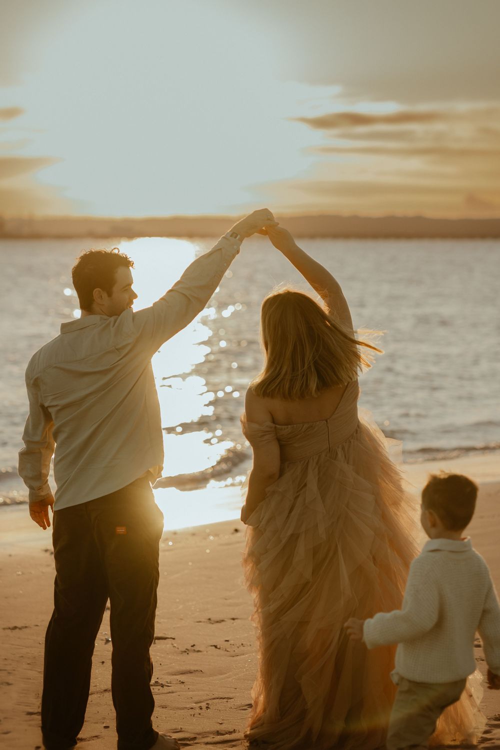 Family silhouettes standing together on beach at golden sunset with light shining through their figures.