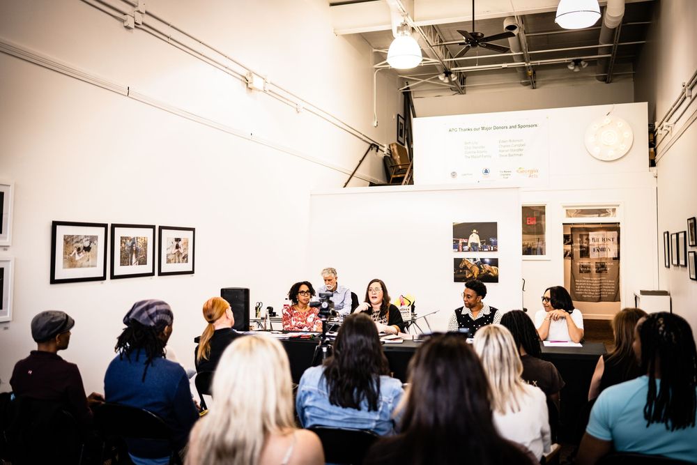 A gallery opening event shows people viewing artwork on white walls under track lighting.