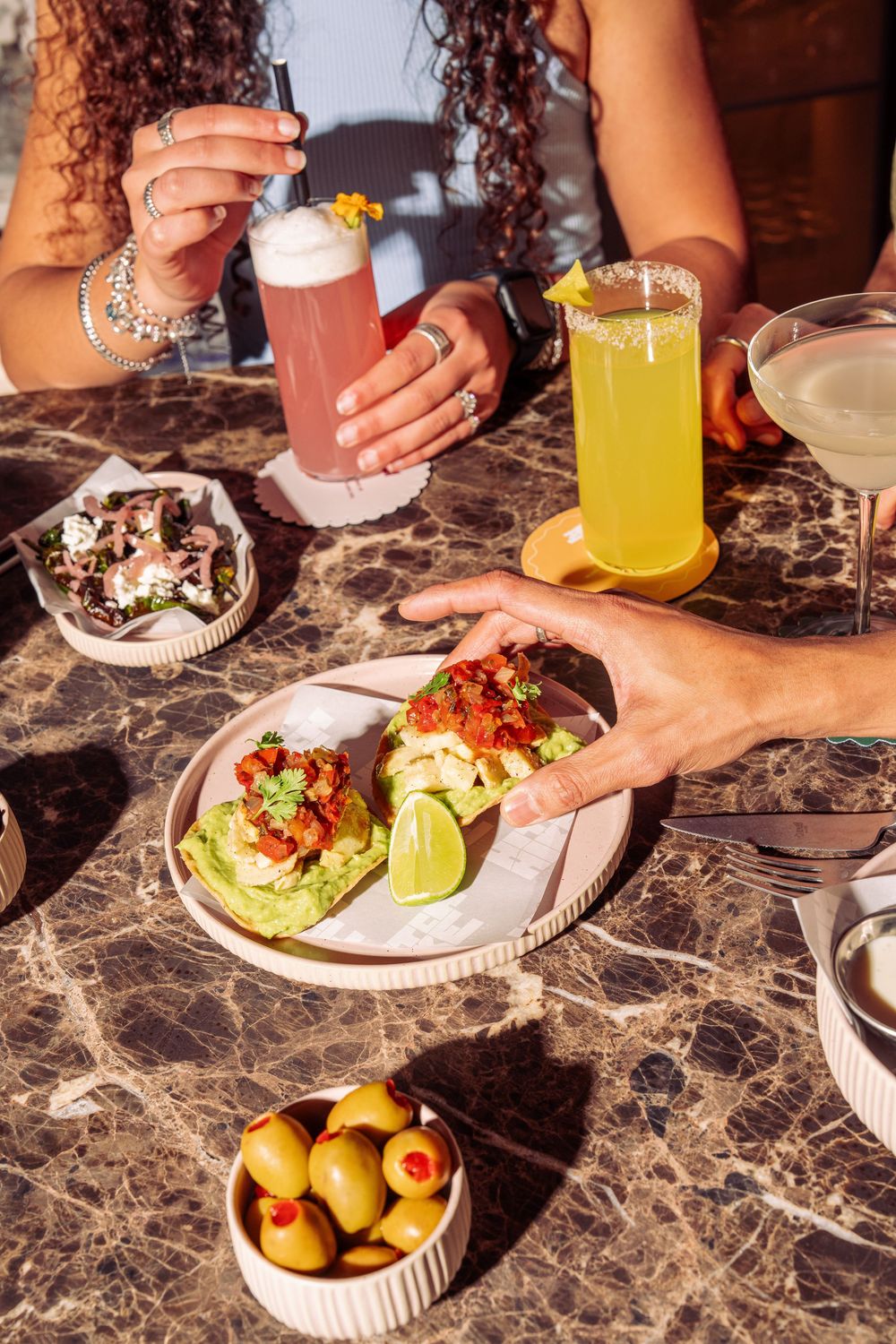 Colorful cocktails and tacos served on marble table with olives and garnishes at a trendy restaurant.