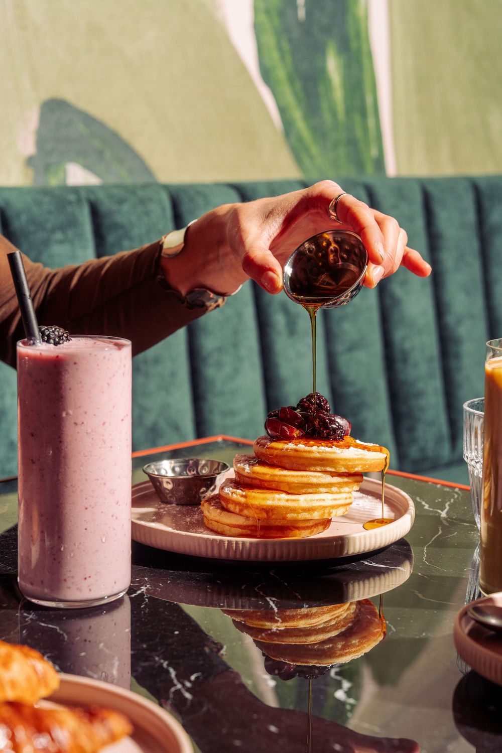 Someone pouring chocolate sauce over a stack of fluffy pancakes with a pink smoothie on a dark table with green seating.
