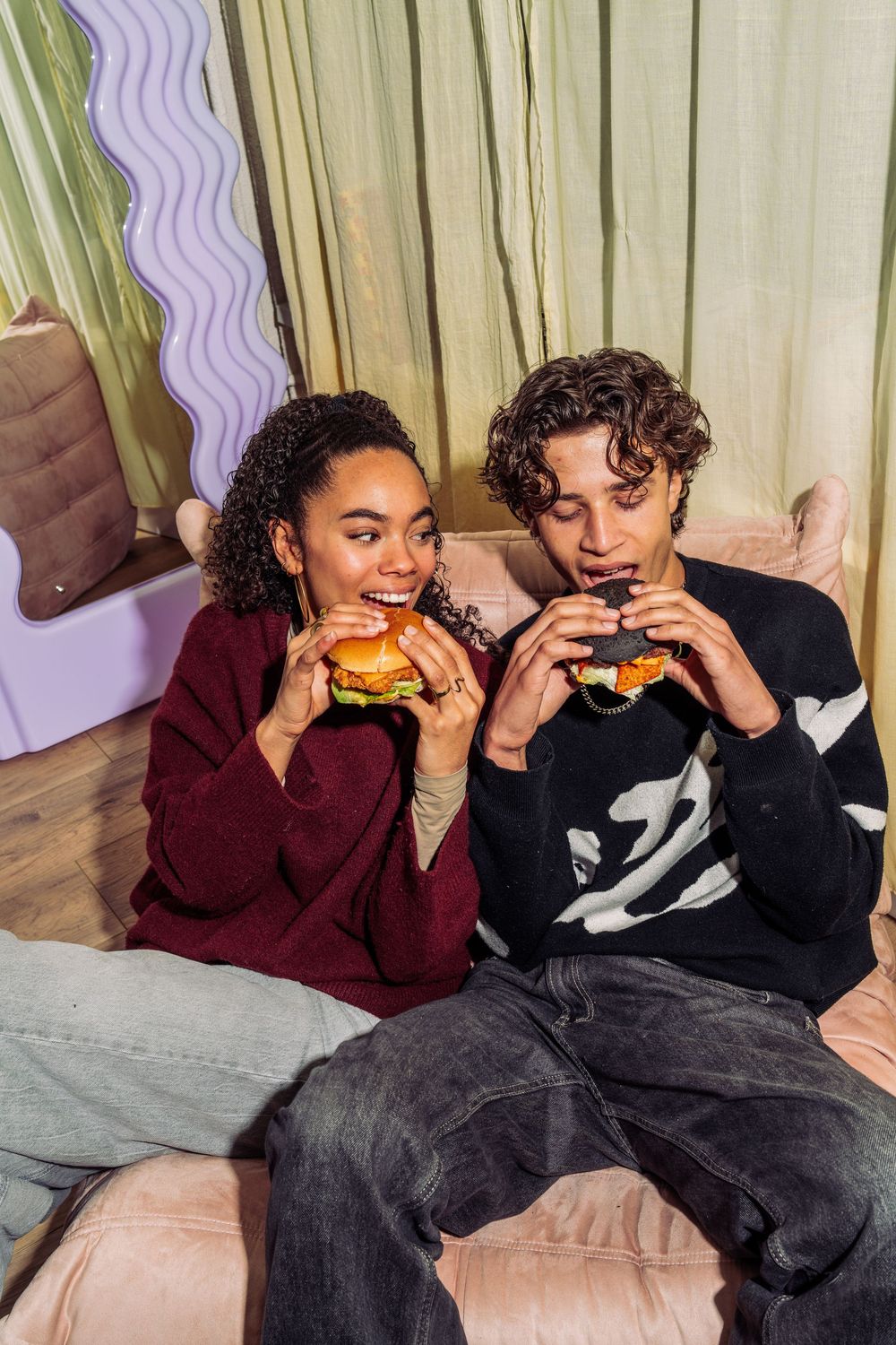 Two people sit together on a couch enjoying burgers in a casual indoor setting.
