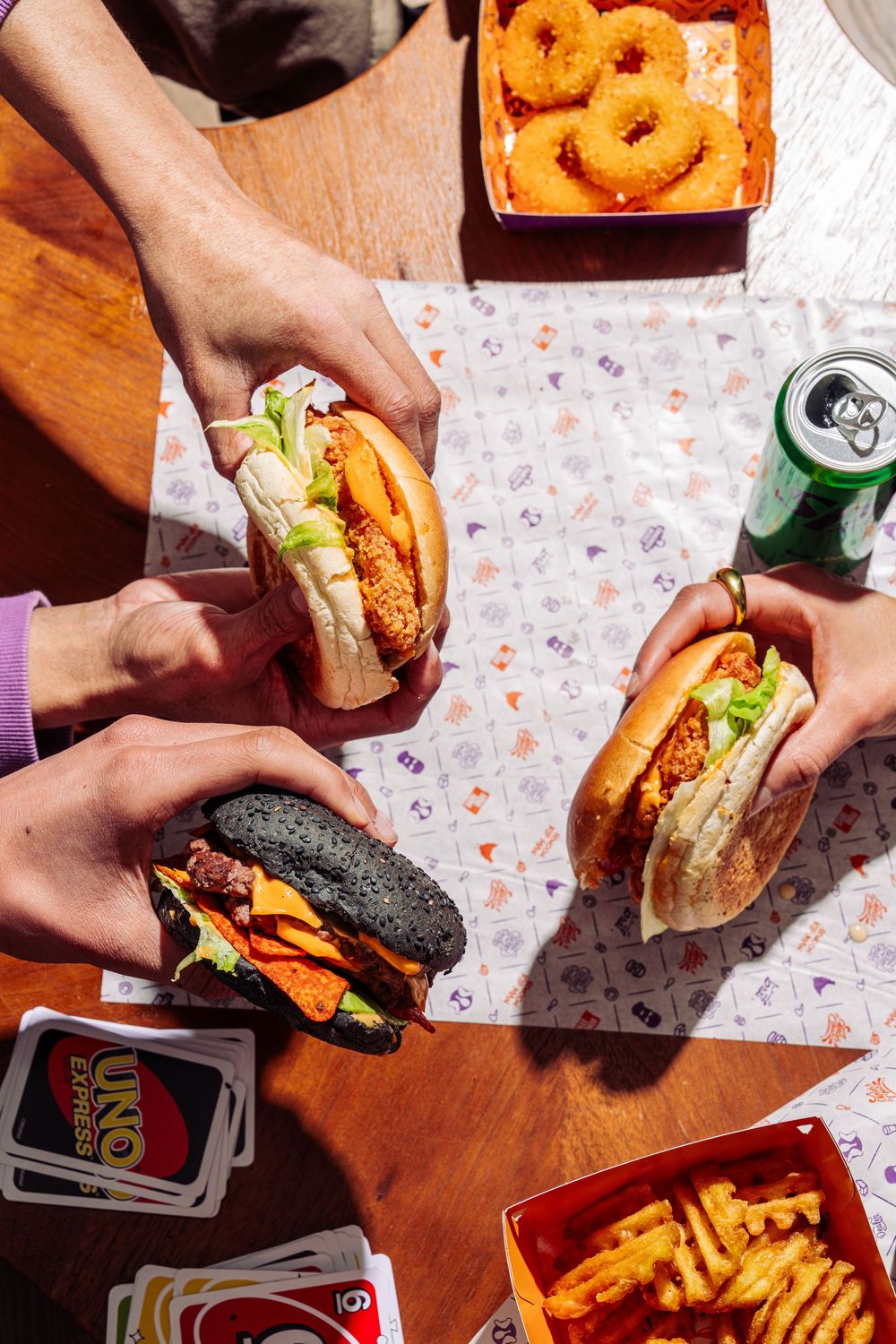 Hands holding burgers and onion rings on a wooden table with colorful paper placemats and french fries.
