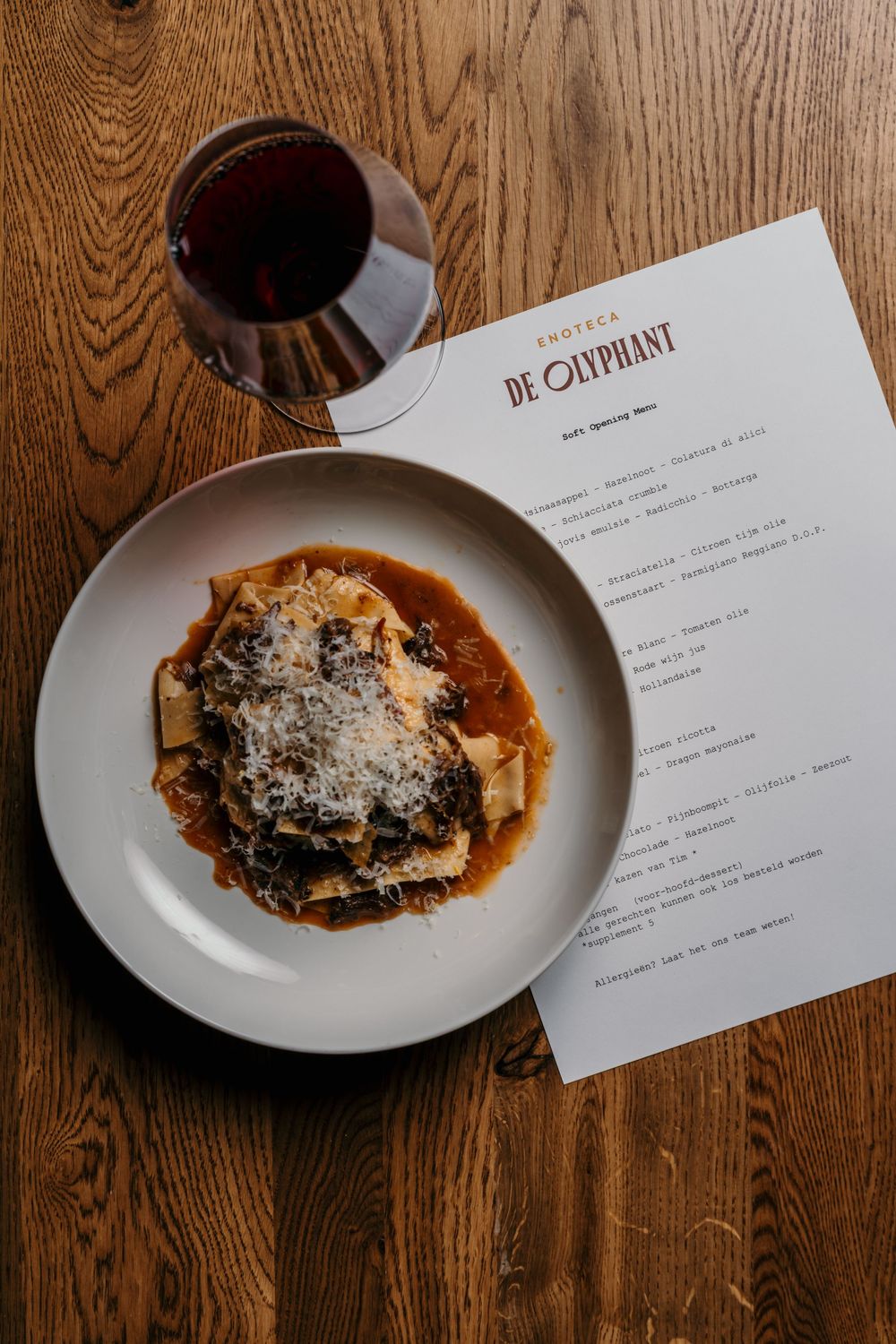 Plate of pasta with sauce and grated cheese next to a wine glass and restaurant menu on wooden table.