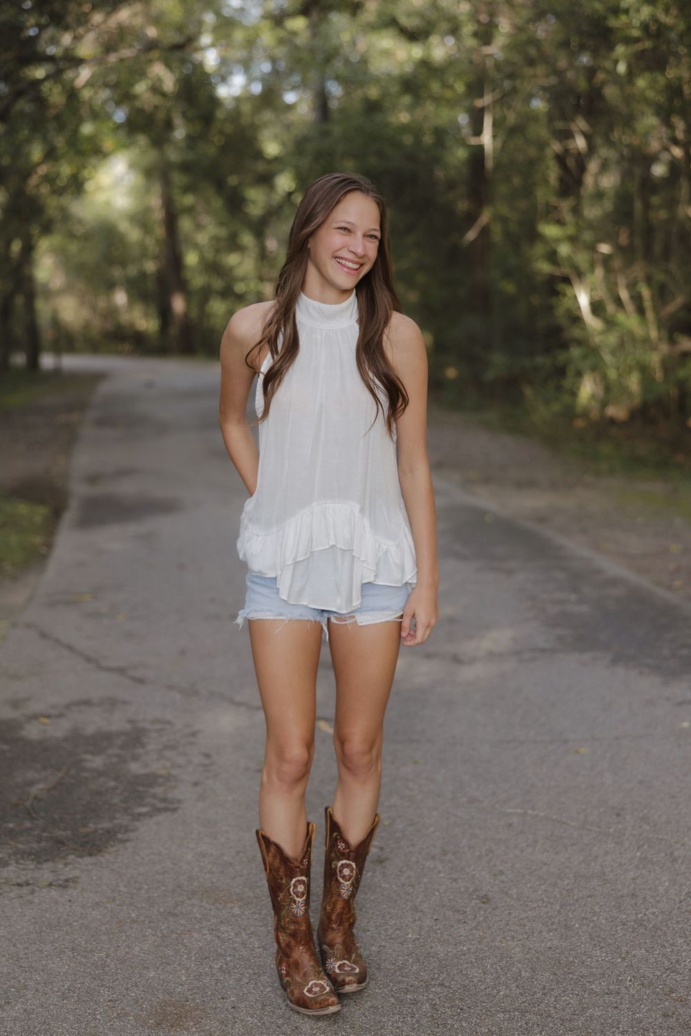 A casual summer outfit featuring a white sleeveless top, denim shorts and brown cowboy boots on a tree-lined path.