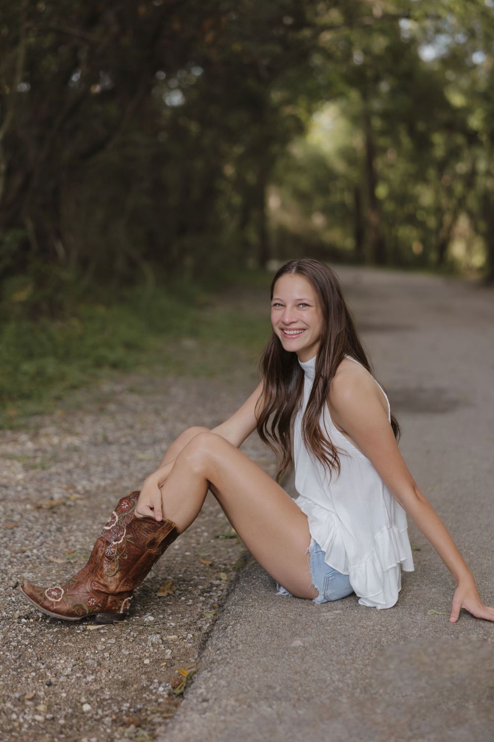 A relaxed outdoor pose showing brown cowboy boots, denim shorts and a flowing white top against a nature backdrop.