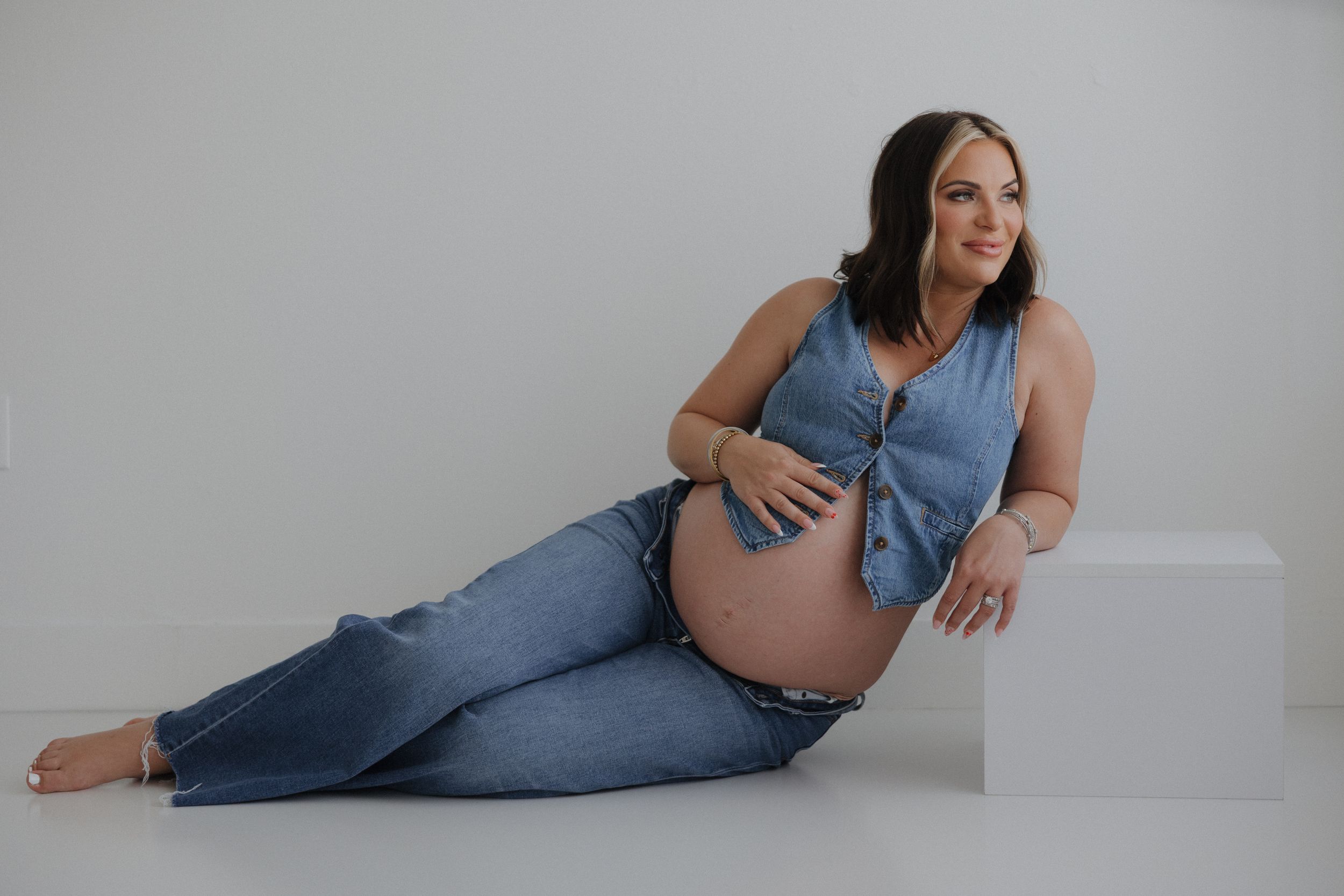 A pregnant woman in denim overalls poses gracefully in a minimalist white studio space during a maternity photoshoot.