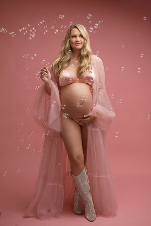 Maternity photo with soap bubbles featuring a pink sheer robe, cowboy boots and pink bikini against coral backdrop.