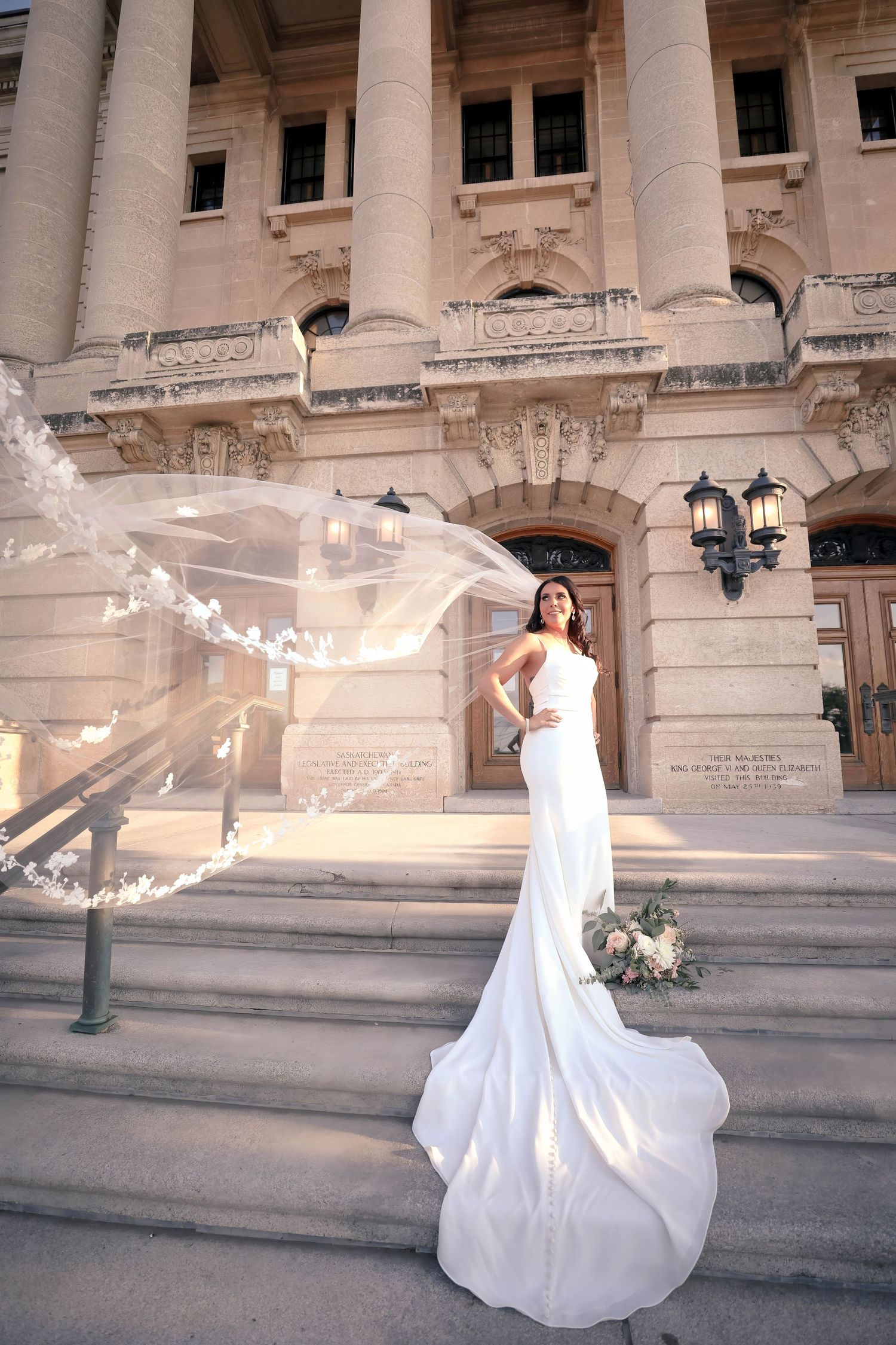 Elegant bride in fitted white gown with flowing veil on grand stone building steps.