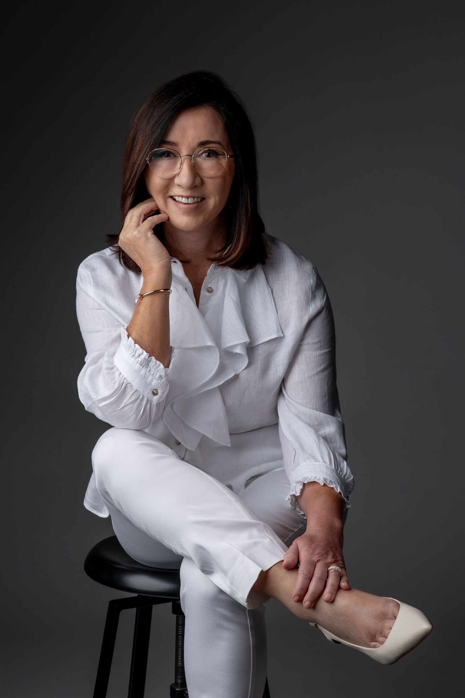 A corporate headshot of a woman in white pants and a blazer, seated comfortably on a stool, smiling warmly against a dark studio background.