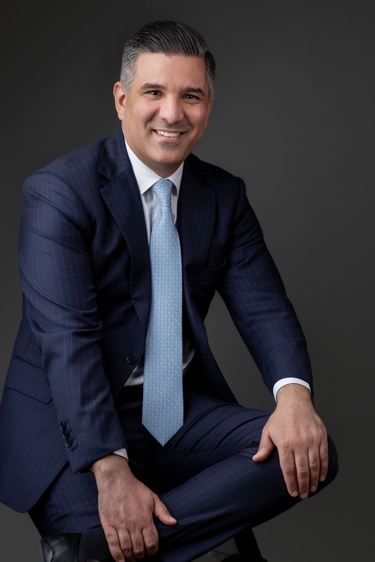A corporate portrait of a man in a navy suit, sitting and smiling confidently against a dark studio background.
