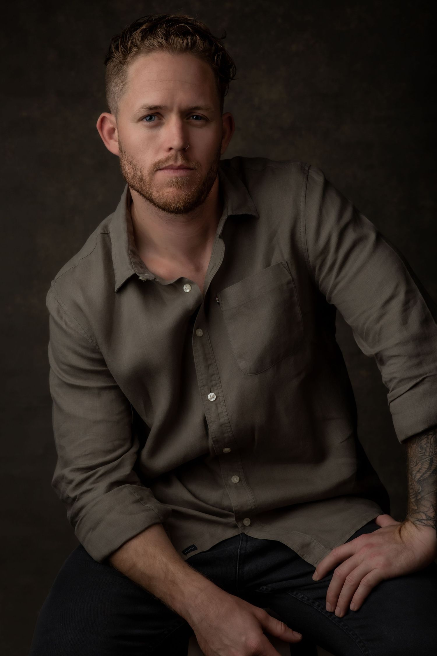 A professional portrait of a man sitting in a dark studio, dressed casually in a button-up shirt, gazing confidently into the camera.