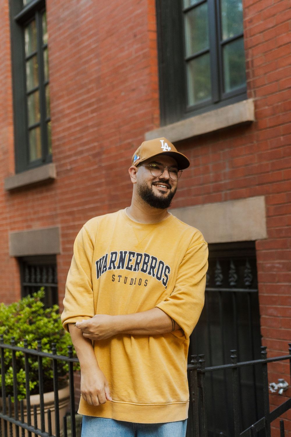 Smiling man in yellow Warner Bros Studios sweatshirt and LA cap poses arms crossed outside red brick building.