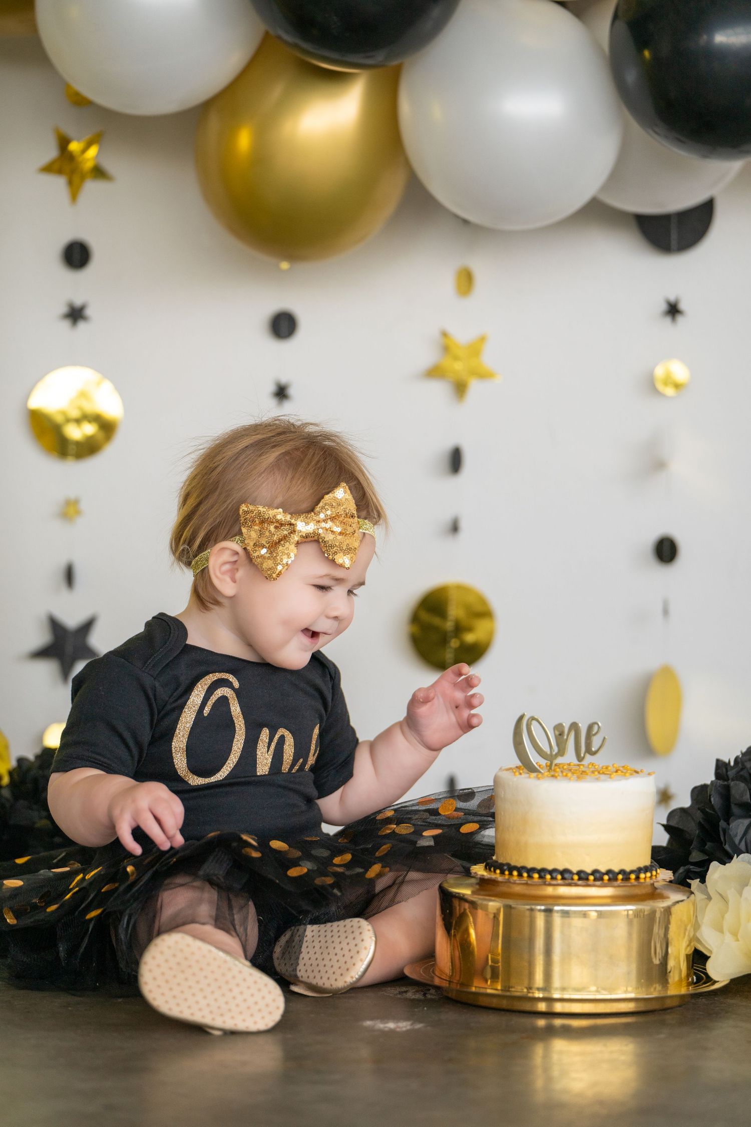 Baby in black and gold outfit celebrating first birthday with cake and balloon decorations.