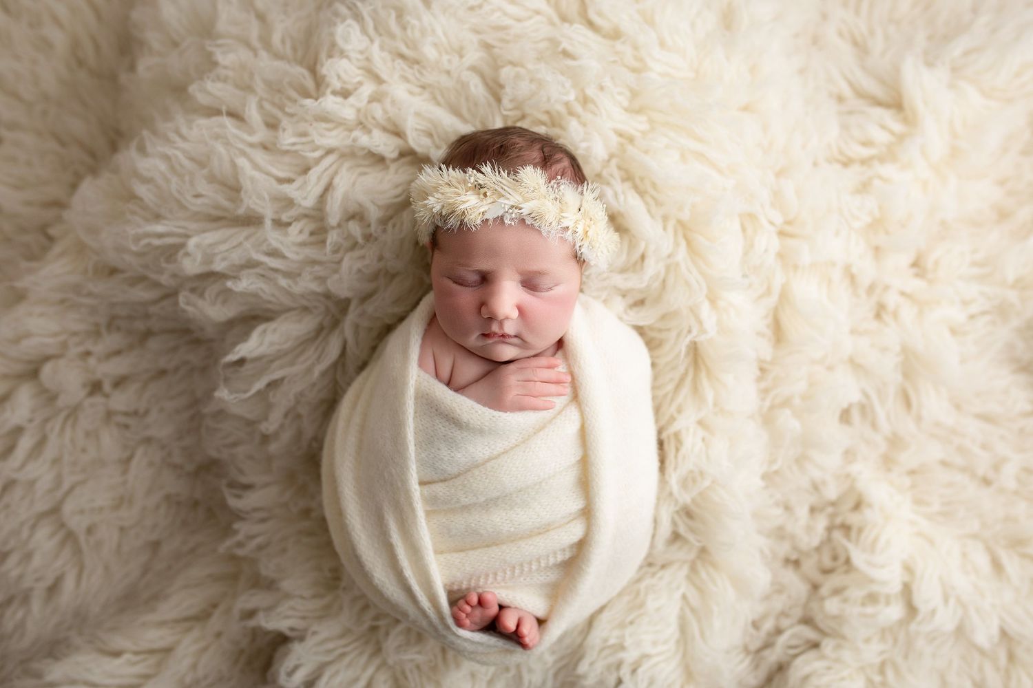 A newborn baby wrapped in white fabric lies peacefully on a soft cream-colored fur blanket in a professional portrait setting.