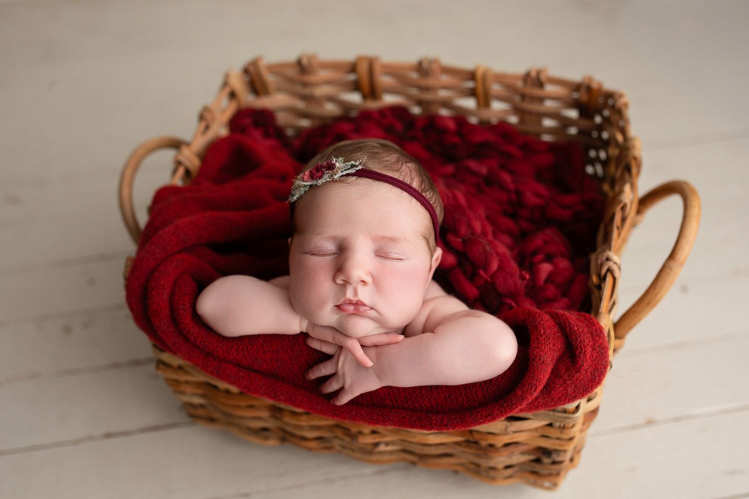 A sleeping newborn nestled in a brown wicker basket with deep red blankets on a white wooden background.