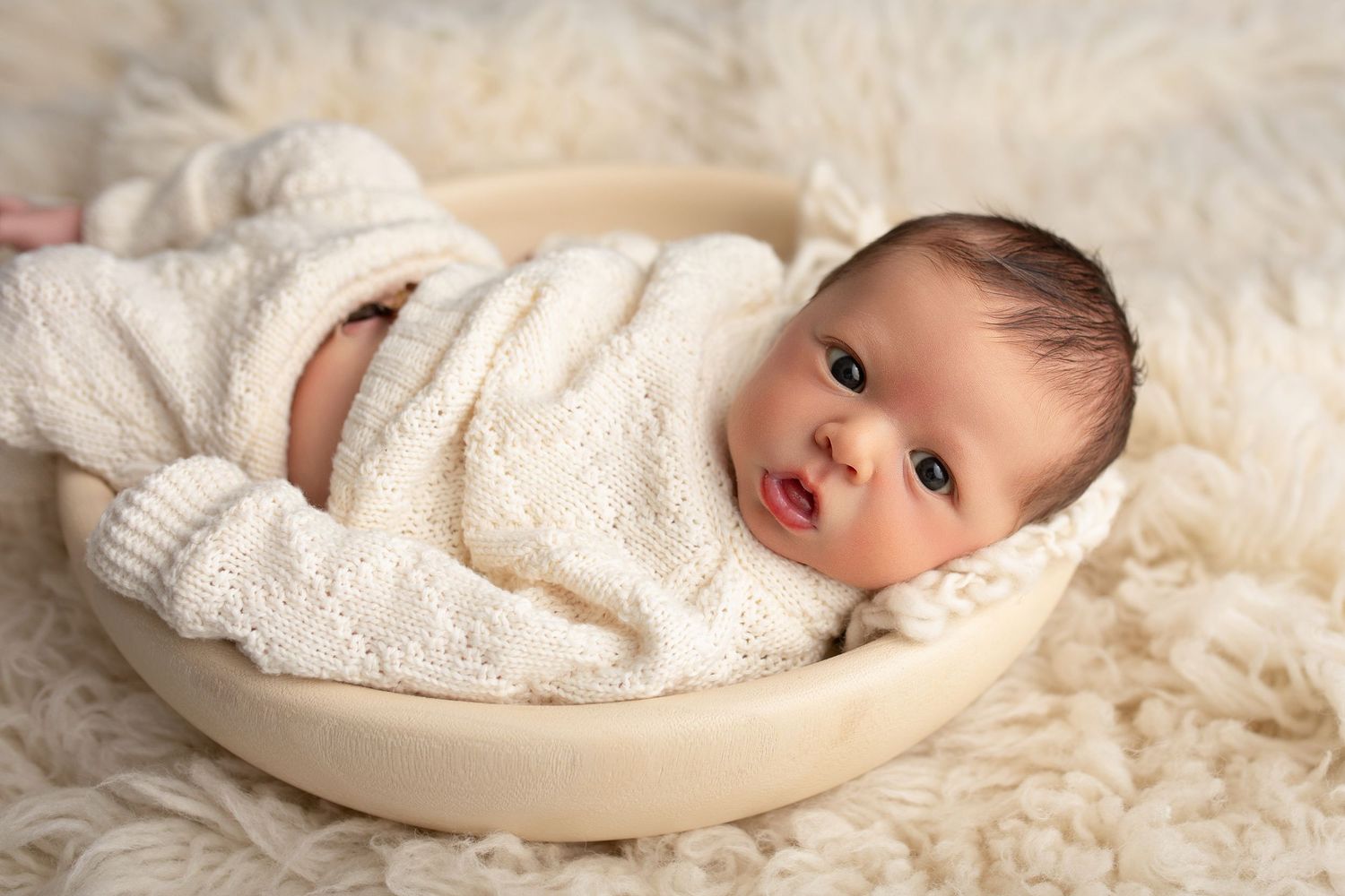 A newborn baby wrapped in soft white blankets rests peacefully on cozy cream-colored fur bedding.