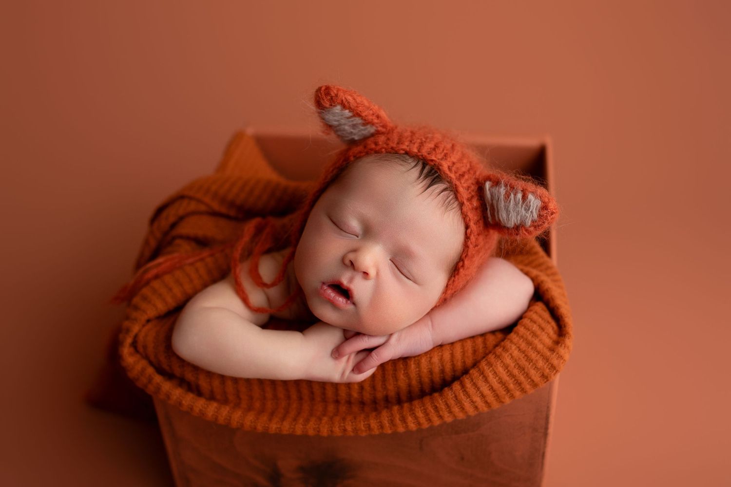 A sleeping newborn baby wearing a coral bow headband nestles in a rust-colored knit blanket.