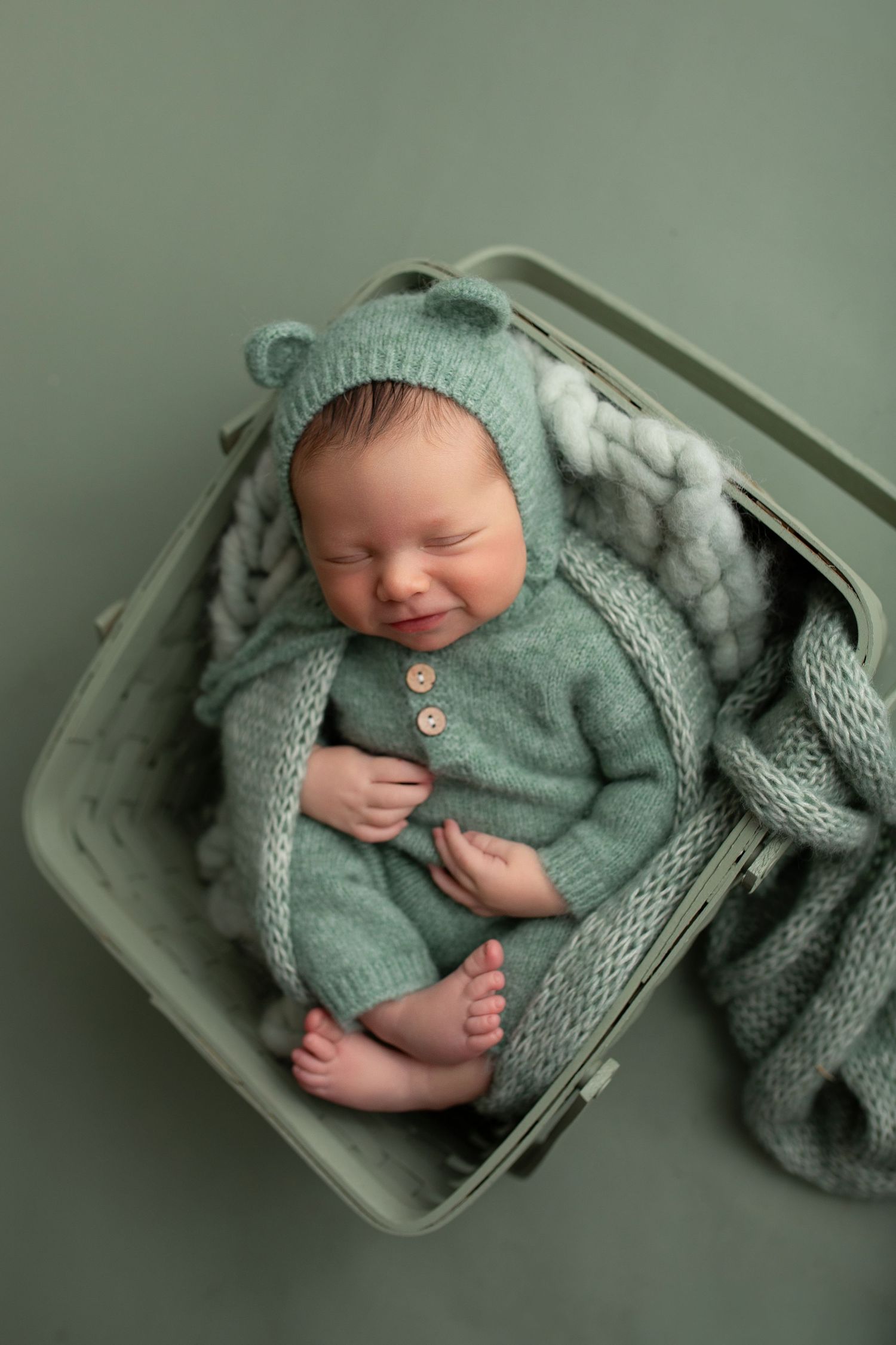 A sleeping newborn in sage green knit wrap lies peacefully in a wooden basket against a matching green backdrop.