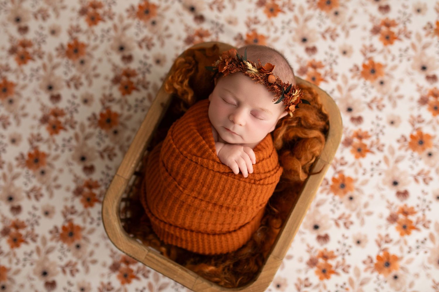 Sleeping newborn baby wearing orange knit outfit rests peacefully in wooden bowl against floral backdrop.