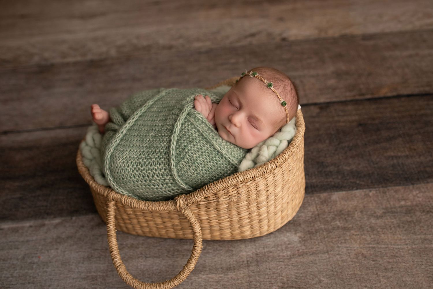 Newborn baby wearing green knit sweater sleeping peacefully in woven basket on rustic wooden floor.
