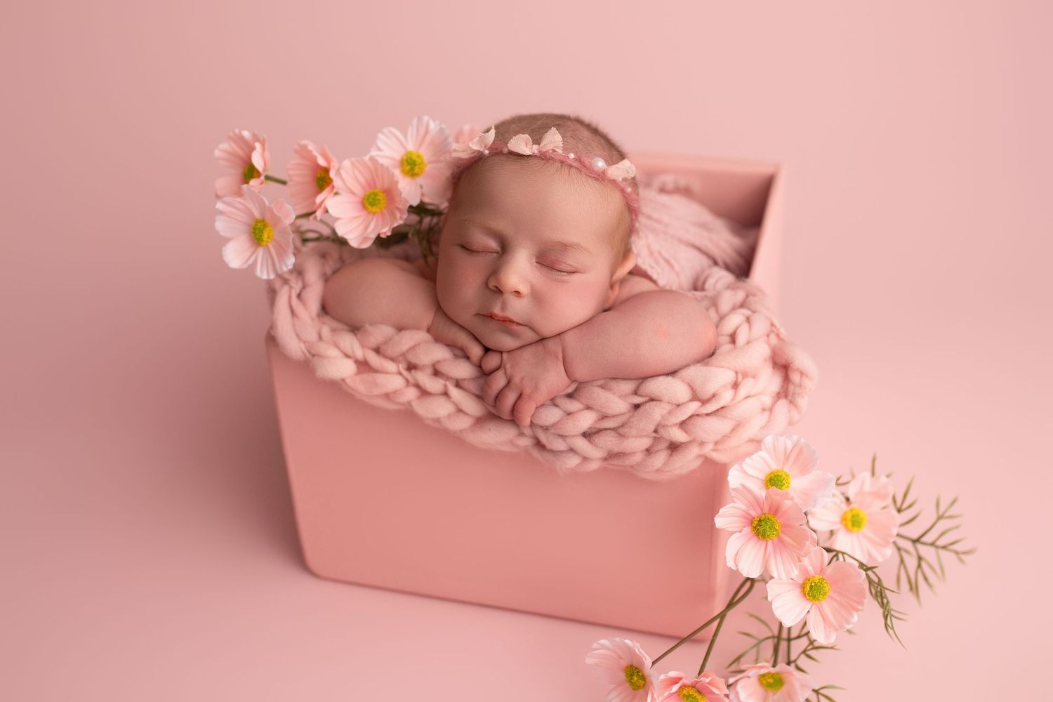 A sleeping newborn baby lies in a pink wooden crate surrounded by delicate pink daisies and soft lighting.
