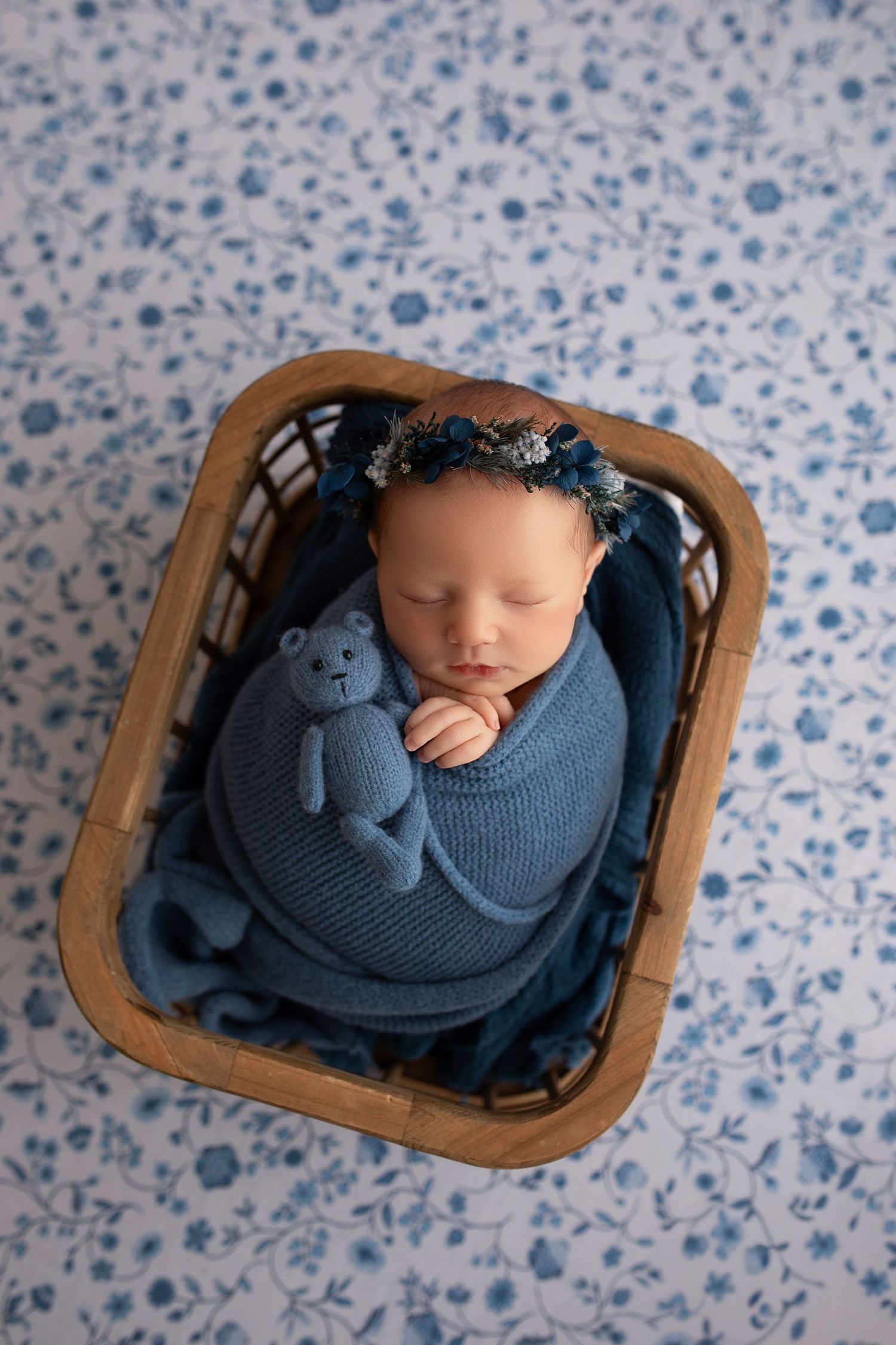 Aerial view of sleeping newborn in blue swaddle and matching decor against blue floral  background.