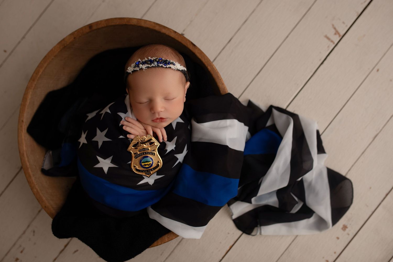 Sleeping newborn wrapped in black, white and blue police-themed blanket in wooden bowl against white wood floor.