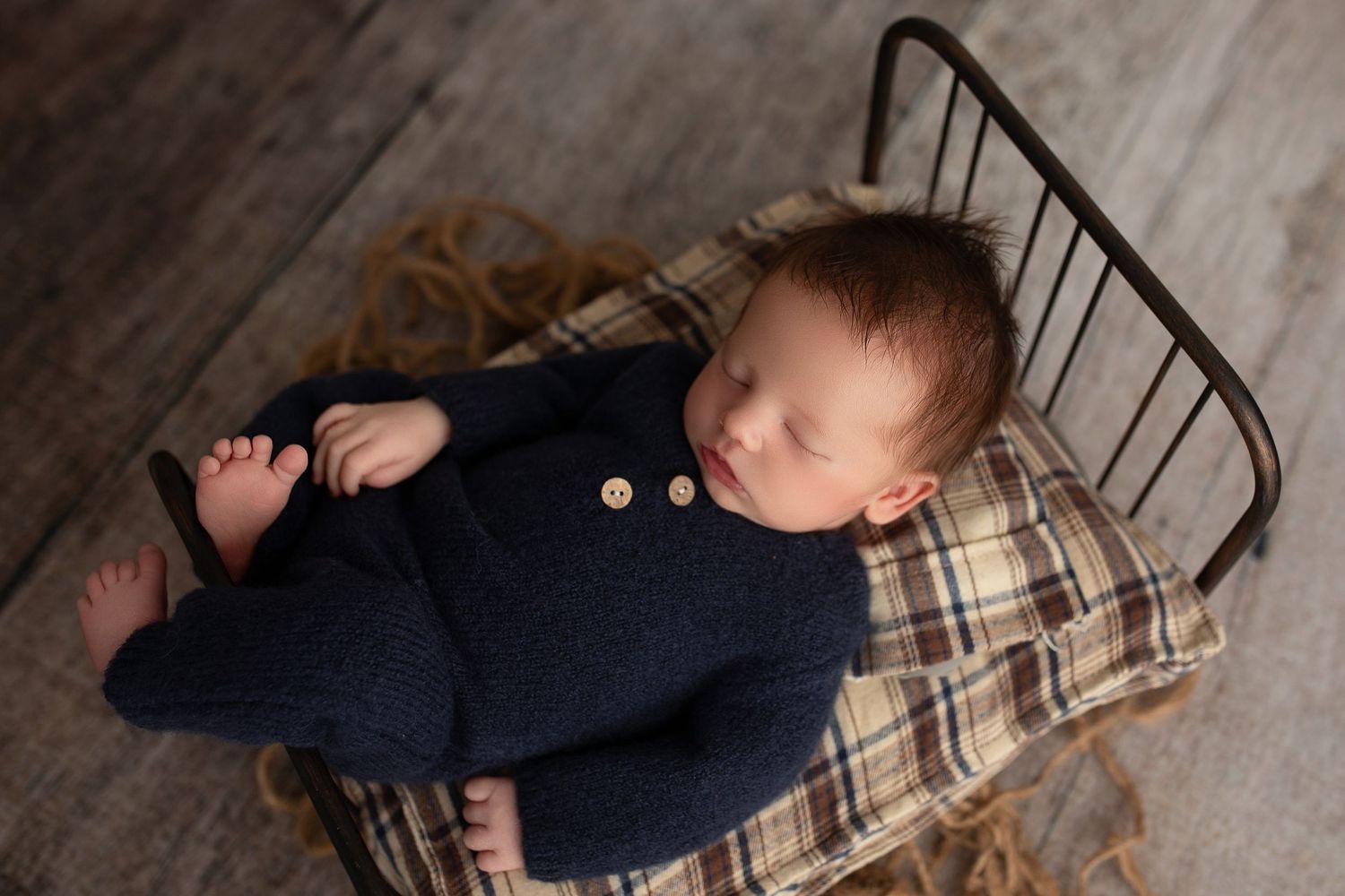 Sleeping baby in navy outfit rests on plaid blanket in vintage metal bed.