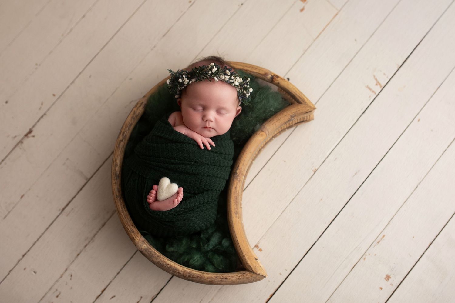 A newborn baby swaddled in black fabric lies in a curved wooden bowl on a white wooden floor.