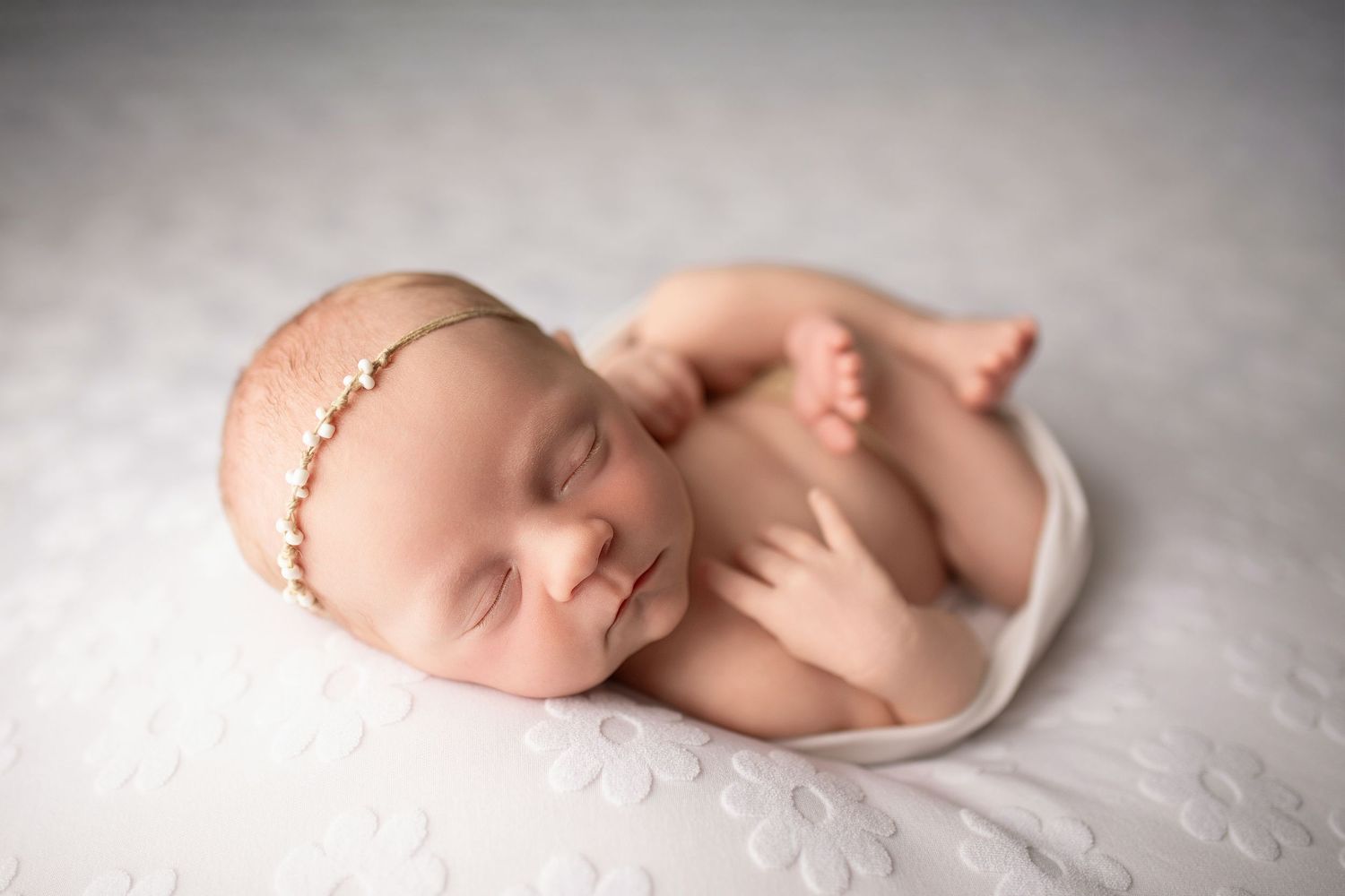 Peaceful newborn baby girl sleeping curled up on white textured blanket wearing a delicate gold floral headband.