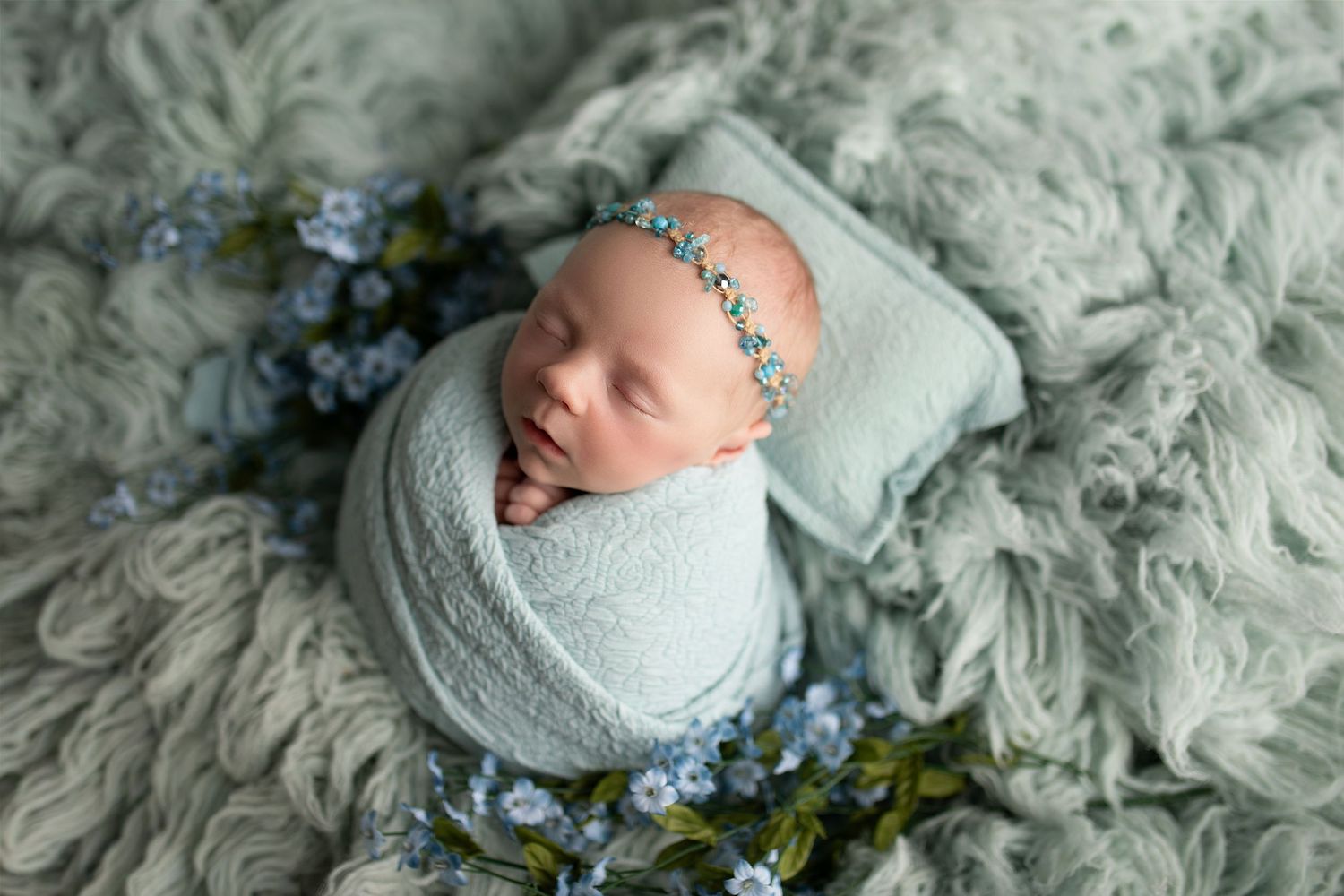 Newborn baby wrapped in mint green swaddle, sleeping peacefully on fluffy blanket with blue flowers and tiny pillow.