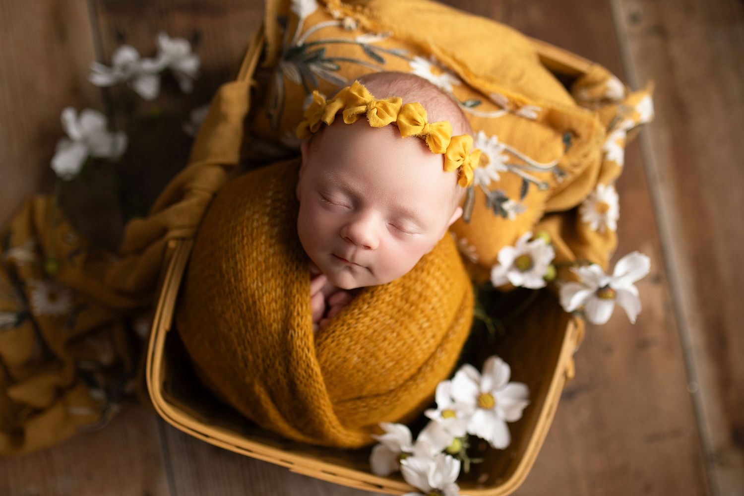 Newborn baby wrapped in mustard yellow knit swaddle with floral headband sleeping in a wicker basket with white flowers.