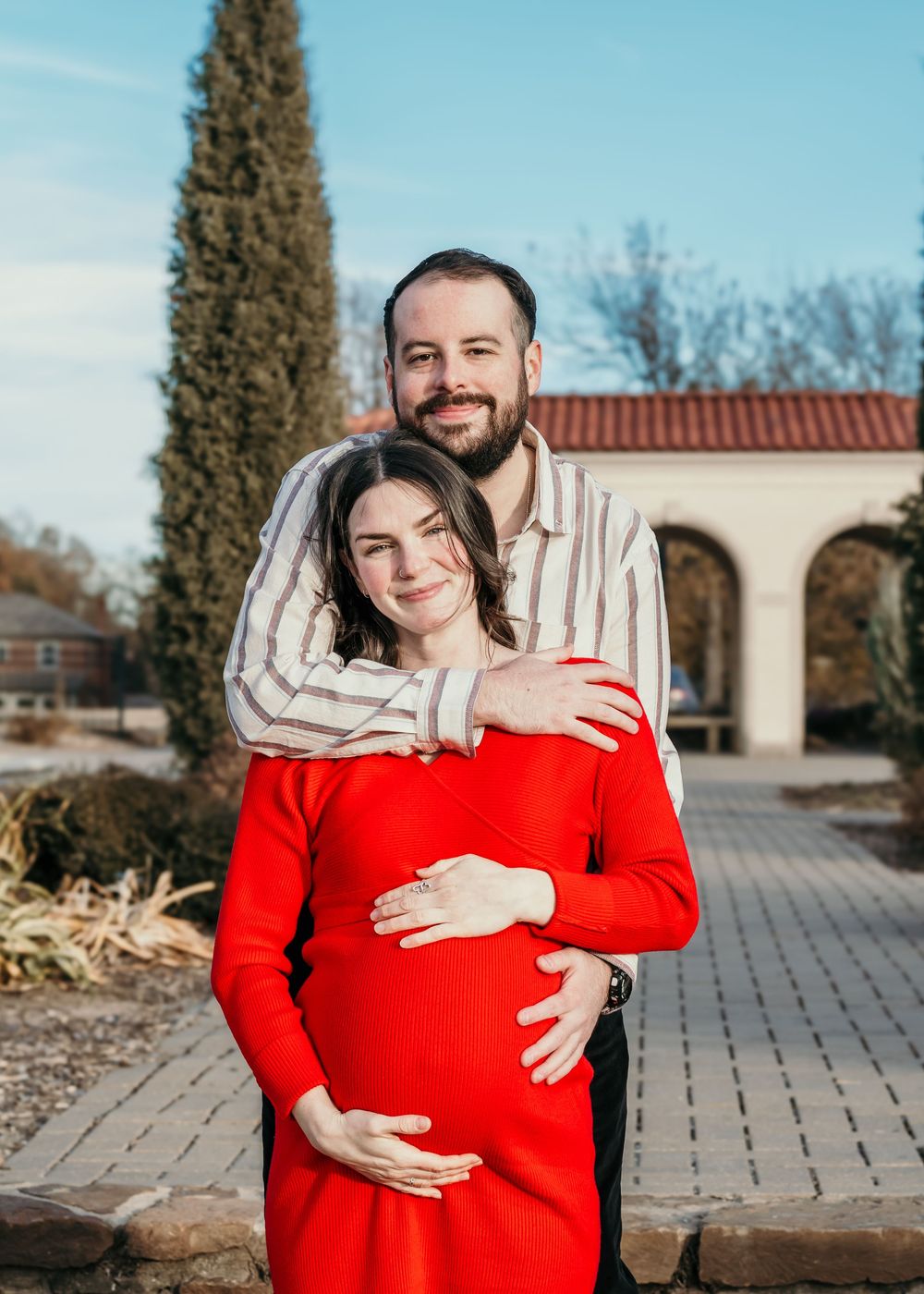 A couple, the man wearing a striped shirt and the woman a red dress, pose in Woodward Park for their maternity session.