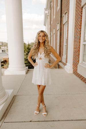 A woman in a short white dress stands confidently in front of a brick building with white columns.