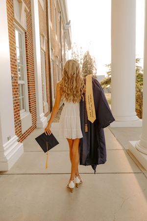 Graduate walks down columned building hallway holding cap and gown in warm sunset lighting.