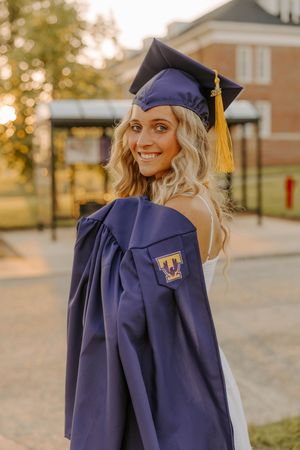 Graduate in navy blue cap and gown smiles over shoulder during sunset photo session.