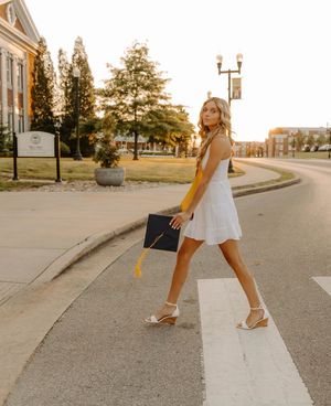 Graduate in white dress walks across campus holding diploma during golden hour.