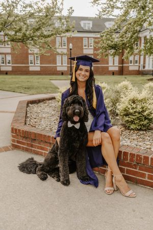 A graduate in purple cap and gown poses with a large black doodle dog on a brick walkway.