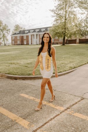 A graduate in white dress and gold sash walking across campus in front of a brick building.
