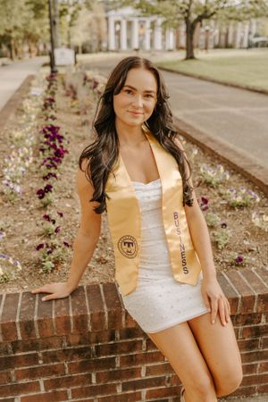 A graduate wearing a gold honors sash poses against a brick wall with flowering garden in background.
