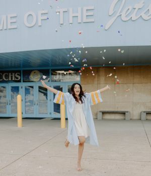 A graduate in white dress and cap celebrating by throwing confetti outside the school building.