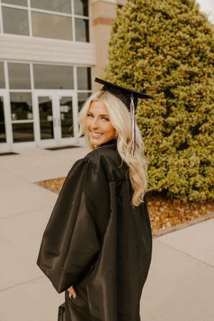 A graduate in black cap and gown poses for a photo outside a school building on graduation day.