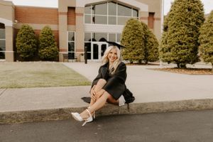 Graduate sits on curb in black graduation gown and cap outside academic building.