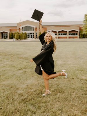 Graduate celebrates by jumping and holding cap in air on grass field near school building.