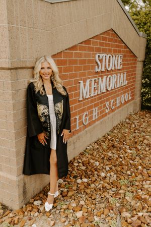 Graduate poses near Stone Memorial High School brick wall sign in traditional black graduation attire.