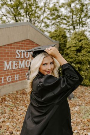 Graduate adjusts cap while posing in front of Stone Memorial school sign wearing black graduation gown.